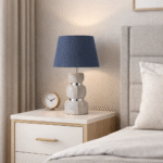 Modern bedside table lamp with blue fabric shade and marble-style base placed on a wooden nightstand next to a bed in a cozy bedroom.